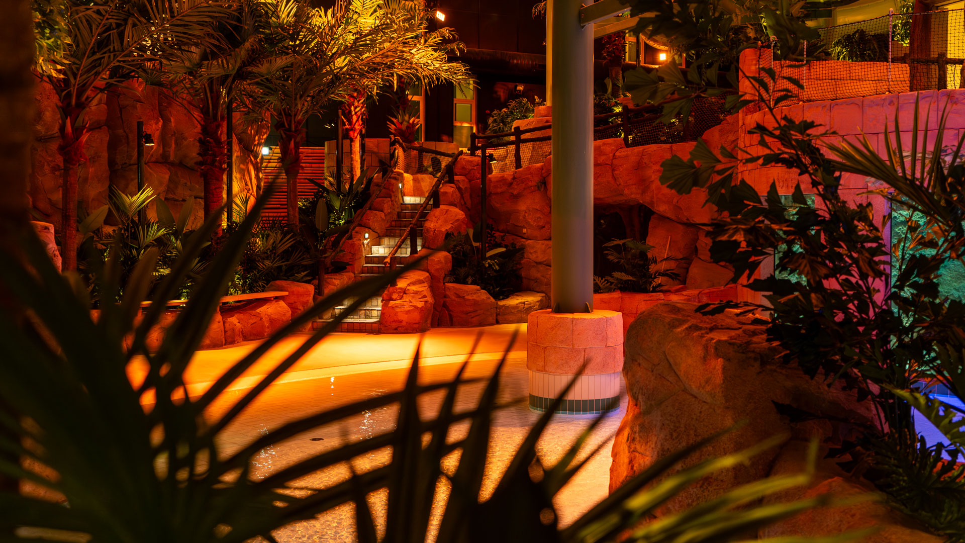A warmly lit indoor tropical pool area features rock formations, lush green plants, palm trees, water, and stairs leading up between rocks, creating a relaxing, resort-like atmosphere.