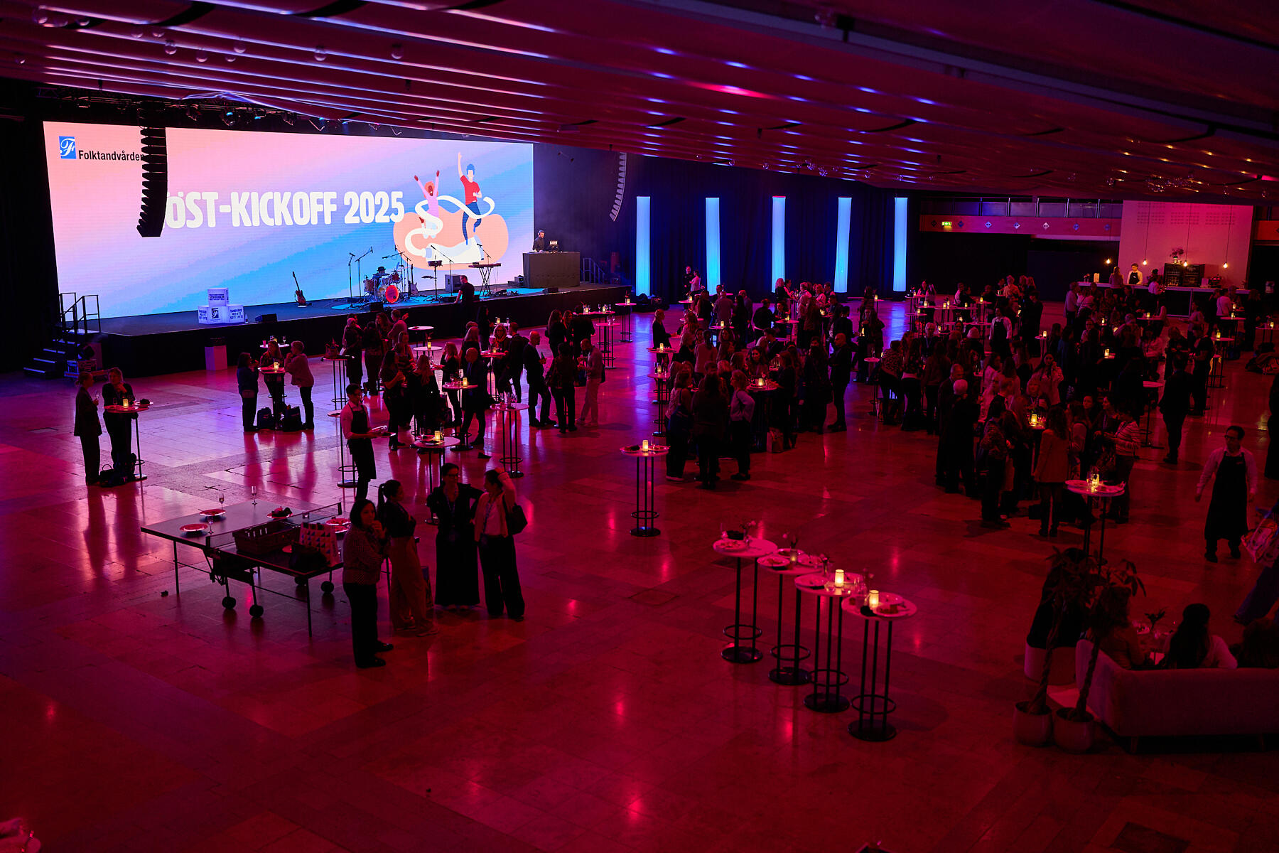 A large indoor event space with groups of people mingling near cocktail tables. A stage with instruments and a large screen displays KICKOFF 2025 in bright letters. The lighting is predominantly pink and purple.