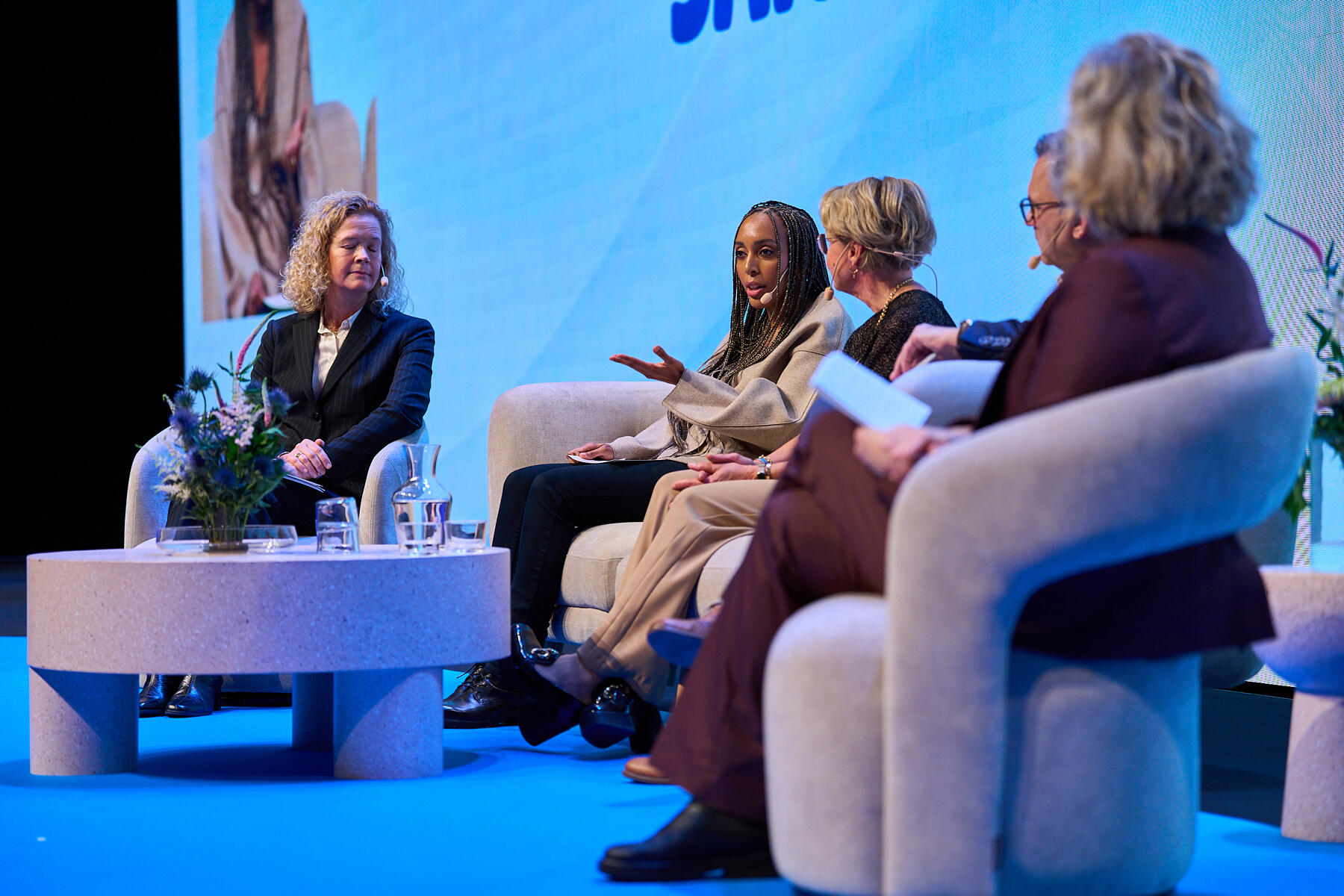 Five people sit in a semi-circle on stage, engaged in a panel discussion. They are seated on modern chairs with small round tables and flowers in front of them. The background is blue with text partially visible.