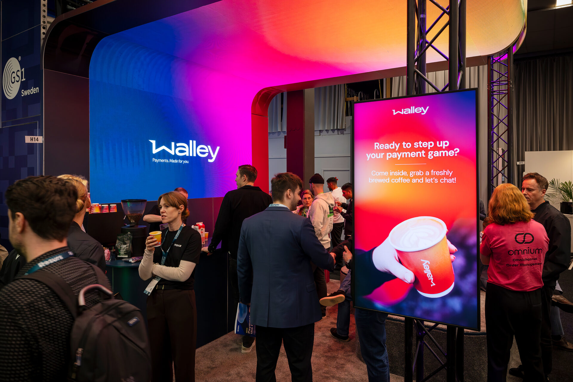 A group of people interact at a tech event booth with colorful lights. A digital sign displays a coffee cup and the text, Ready to step up your payment game? Come inside, grab a freshly brewed coffee and lets chat!.
