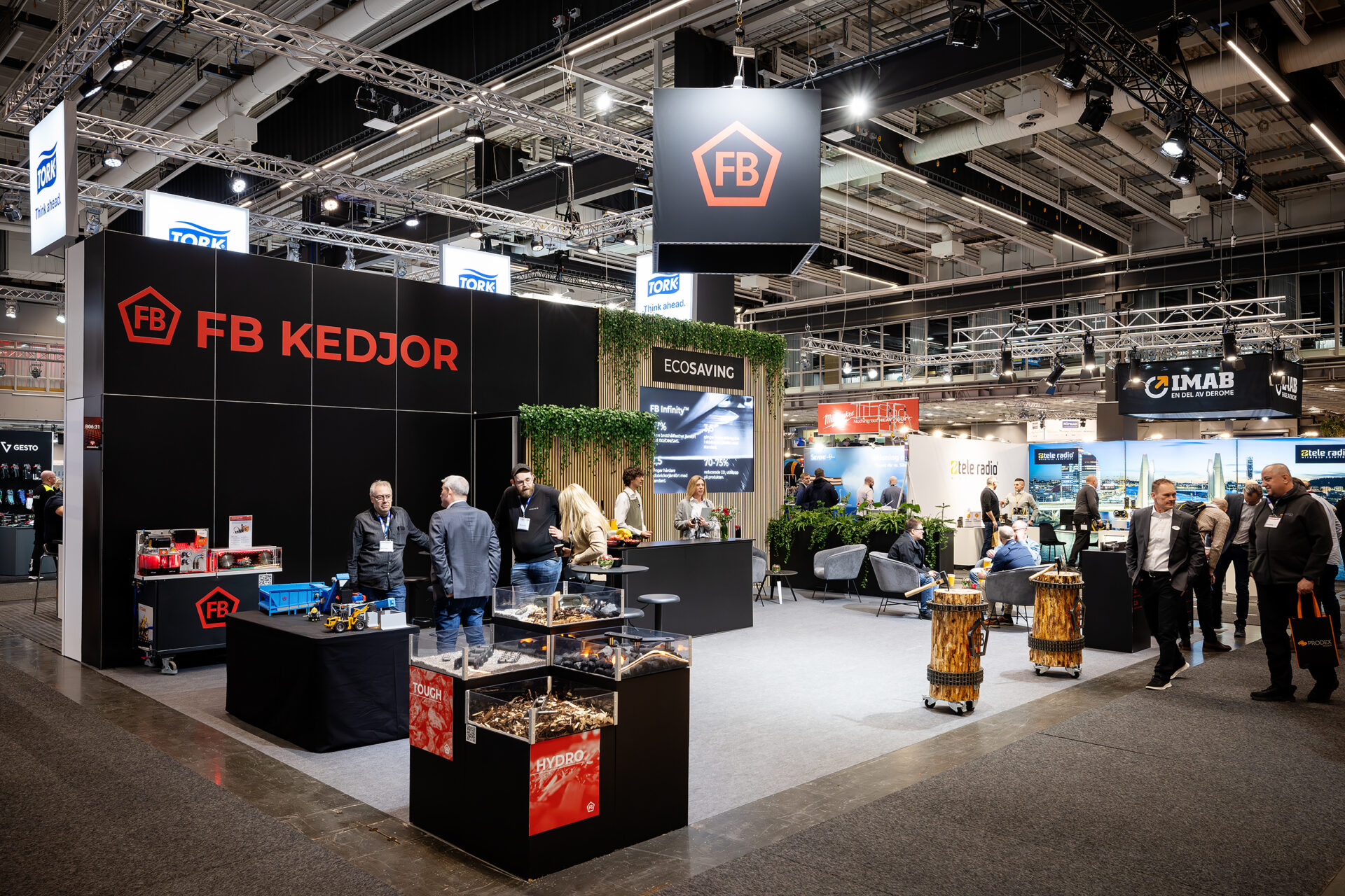 A trade show booth for FB Kedjor features people conversing, product displays, and branded signage. Other booths and attendees are visible in a large, well-lit exhibition hall.