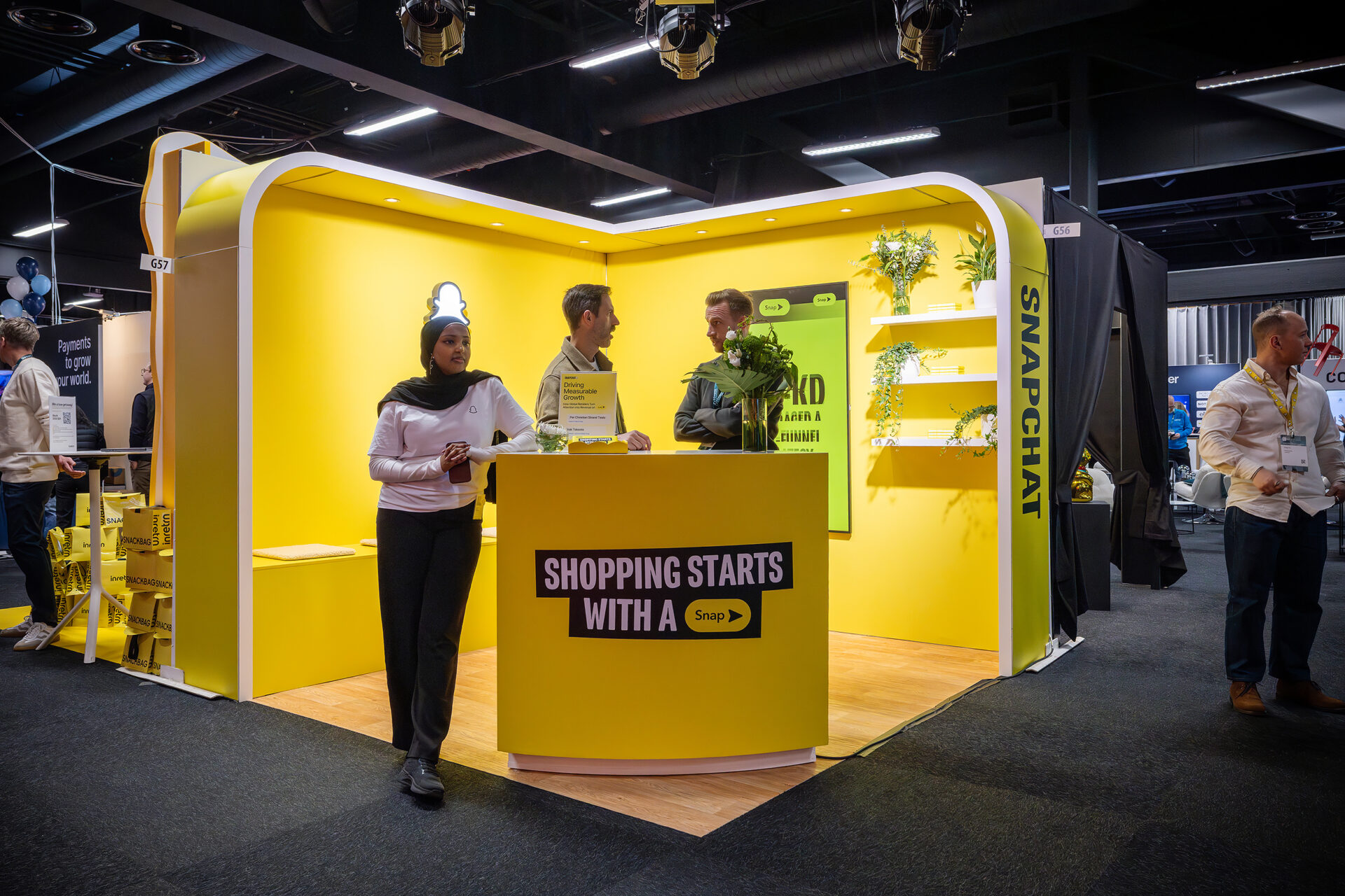 A bright yellow Snapchat booth at an event, with three people interacting. A woman stands in front holding a phone, while two men inside the booth converse near a counter that reads “Shopping starts with a Snap.”.