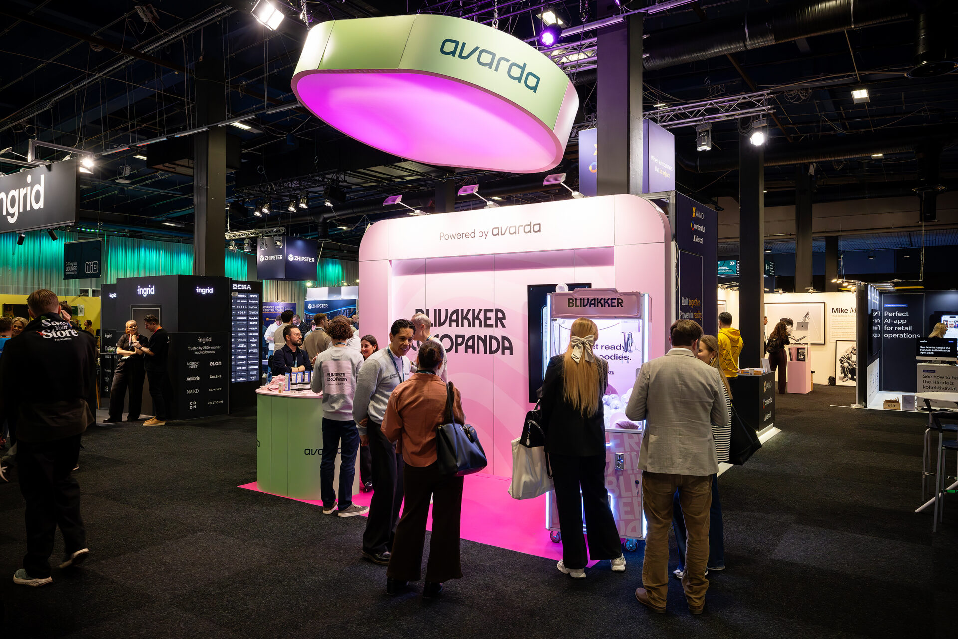 People gather around a brightly lit exhibition booth with a large oval green and pink sign reading avarda at a tech event. Other booths and attendees are visible in the background under dark lighting.