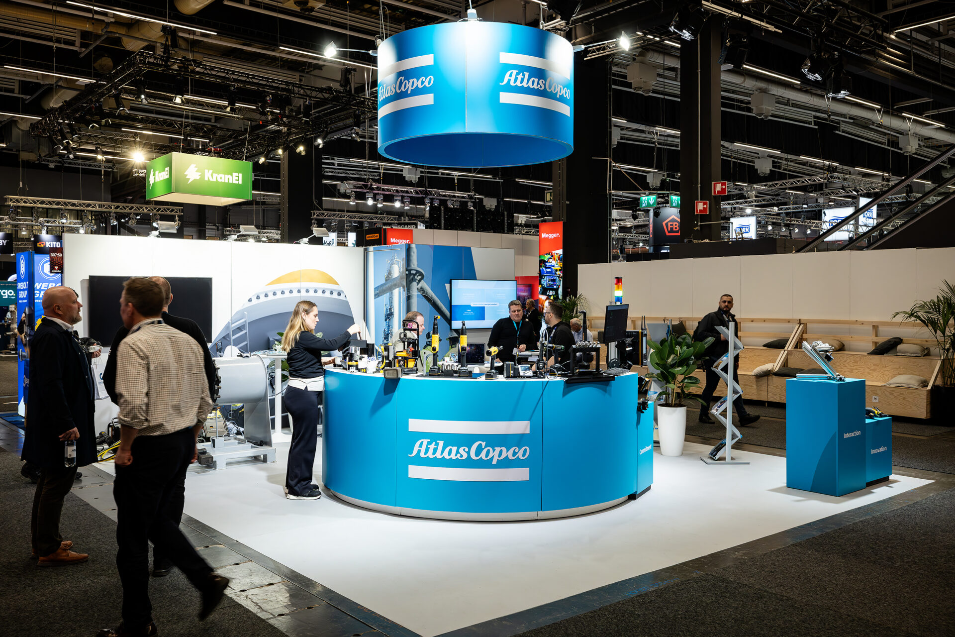 A brightly lit trade show booth for Atlas Copco features a circular blue counter, branded signs, product displays, and staff engaging with visitors on a busy exhibition floor.