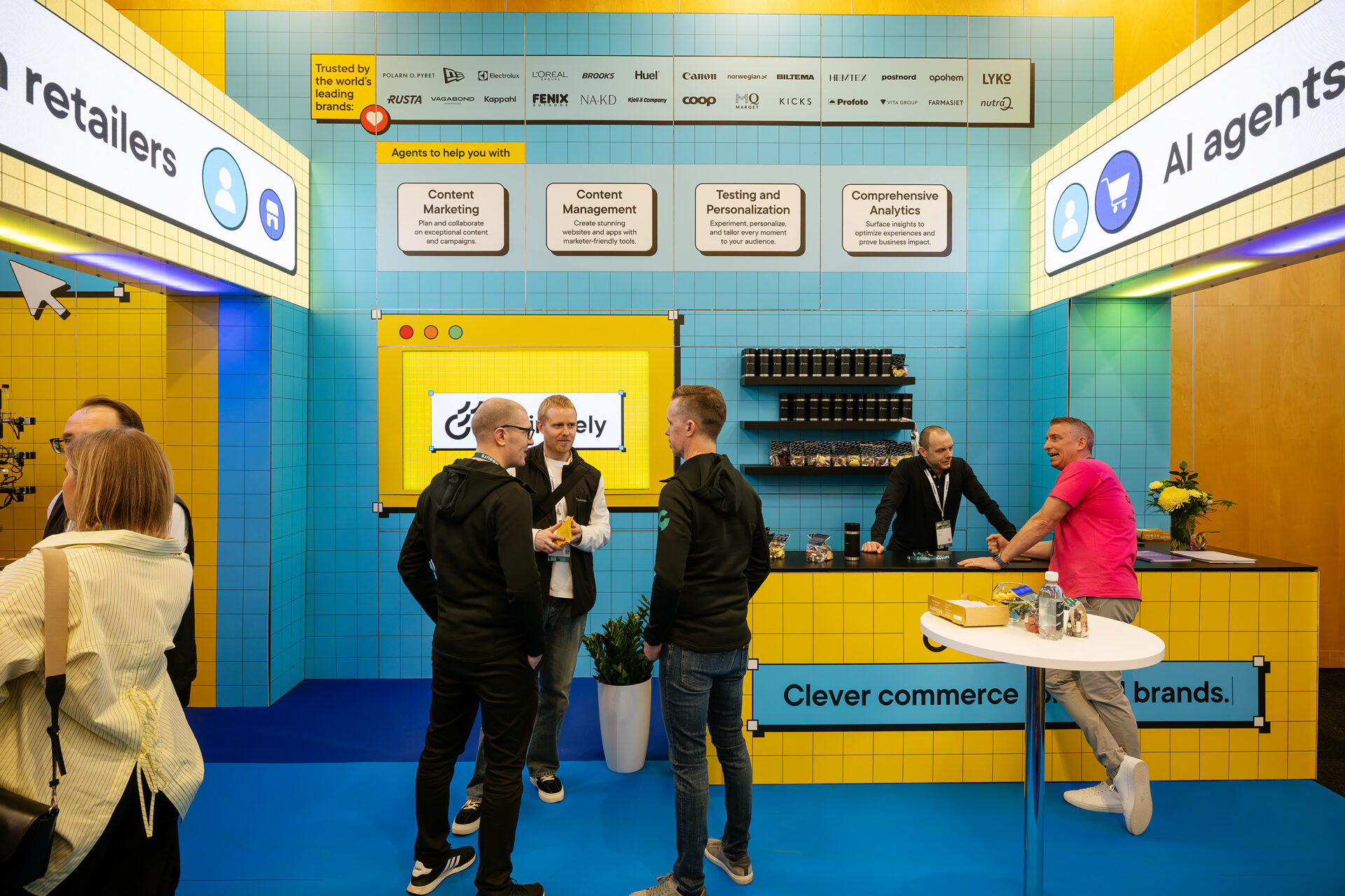 A group of people talk and network at a brightly colored trade show booth with yellow and blue walls, featuring banners about AI agents and commerce, plants, and a counter with drinks and snacks.
