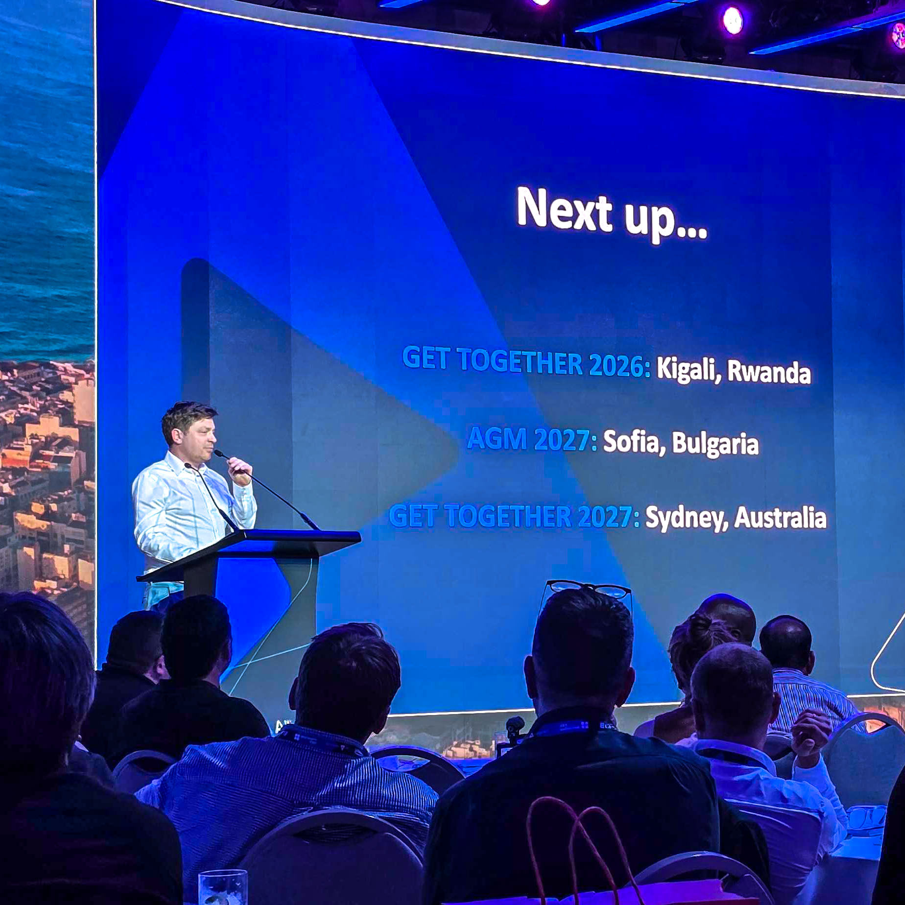 A speaker stands at a podium addressing an audience. A large screen behind him displays upcoming event locations: Kigali, Rwanda (2026), Sofia, Bulgaria (2027), and Sydney, Australia (2027).