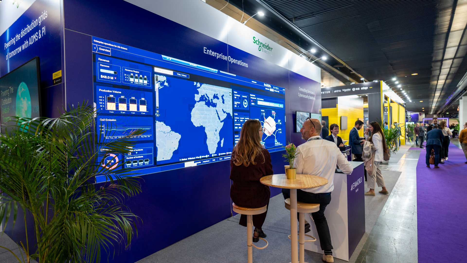 People interact with a large digital display showing global data at a Schneider Electric booth during a trade show, with other booths and attendees visible in the brightly lit exhibition hall.